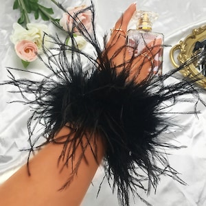 May include: A black feather wrist cuff. The cuff is made of a thick layer of black feathers that are arranged in a circular shape. The cuff is worn on a person's wrist.