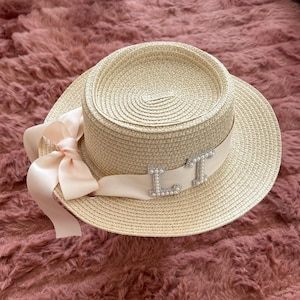Può includere: Un cappello di paglia con un nastro bianco e le lettere "LT" decorate con perle sul nastro. Il cappello è su una superficie rosa pelosa.