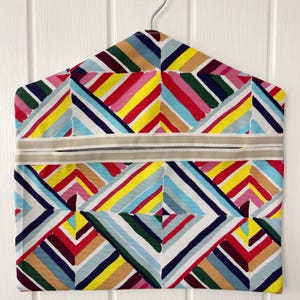 Pode incluir: Uma colorida capa para roupas com um padrão geométrico de listras diagonais em vermelho, amarelo, azul e verde. A capa tem uma faixa bege no meio e pendura em um cabide de metal. Projetada para proteger roupas.