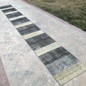 May include: A long, rectangular rug with a patchwork design in shades of gray, white, and green. The rug is laid out on a gray concrete walkway.