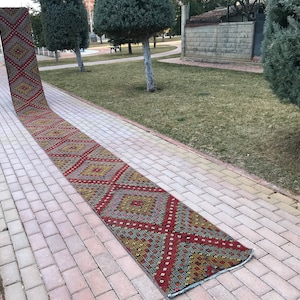 May include: A long, narrow, red, yellow, and blue patterned rug laid out on a brick walkway. The rug has a geometric design with diamonds and squares.
