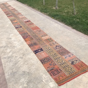 Könnte beinhalten: Ein langer, rechteckiger Vintage-Teppich mit Patchwork-Design. Der Teppich weist eine Vielzahl von Farben auf, darunter Orange, Rot, Grün und Blau. Der Teppich liegt auf einer Betonoberfläche, wobei auf beiden Seiten Gras zu sehen ist.
