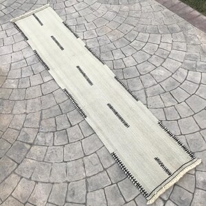 May include: A long, off-white runner rug with black stitching and fringe. The rug has a simple geometric pattern.