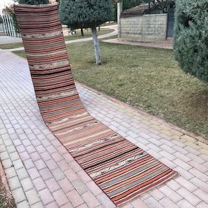 May include: A long, narrow, handwoven rug with a striped pattern in shades of red, orange, brown, and beige. The rug is laid out on a brick walkway.