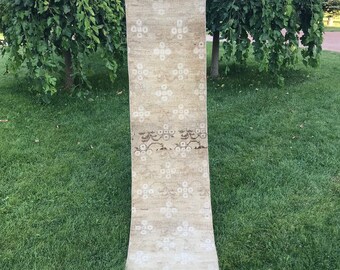 May include: A long, beige runner rug with a white floral pattern. The rug is folded in half and held by two hands.