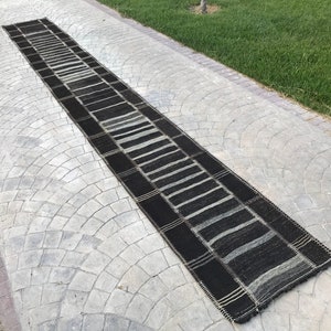 May include: A long, narrow, rectangular rug with a striped pattern in shades of black, gray, and white. The rug is placed on a gray concrete surface, with green grass visible in the background. The rug's design features alternating dark and light stripes.