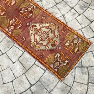 May include: A vintage Turkish runner rug with a geometric pattern in shades of red, orange, and brown. The rug has a fringed edge and is laid out on a stone floor.