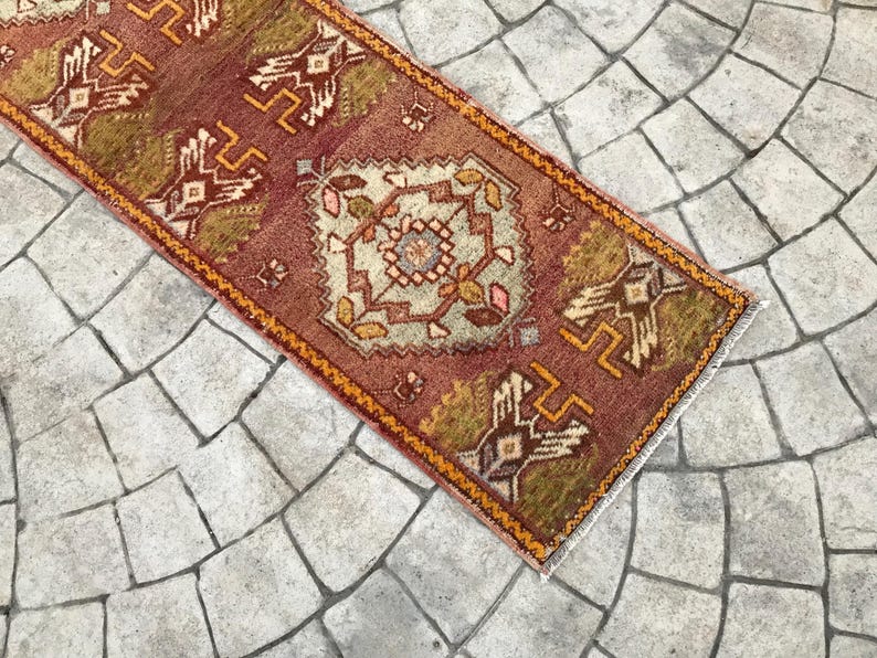 May include: A vintage Turkish runner rug with a geometric pattern in shades of red, orange, and brown. The rug has a fringed edge and is laid out on a stone floor.