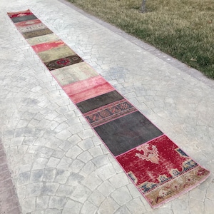 May include: A long, rectangular patchwork rug with a variety of colours and patterns. The rug features sections of red, pink, grey, and beige. The rug is placed on a grey, textured surface, with grass visible in the background.