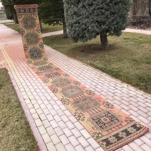 May include: A long, narrow, vintage rug with a colorful geometric pattern in shades of pink, orange, blue, and black. The rug is laid out on a brick walkway.