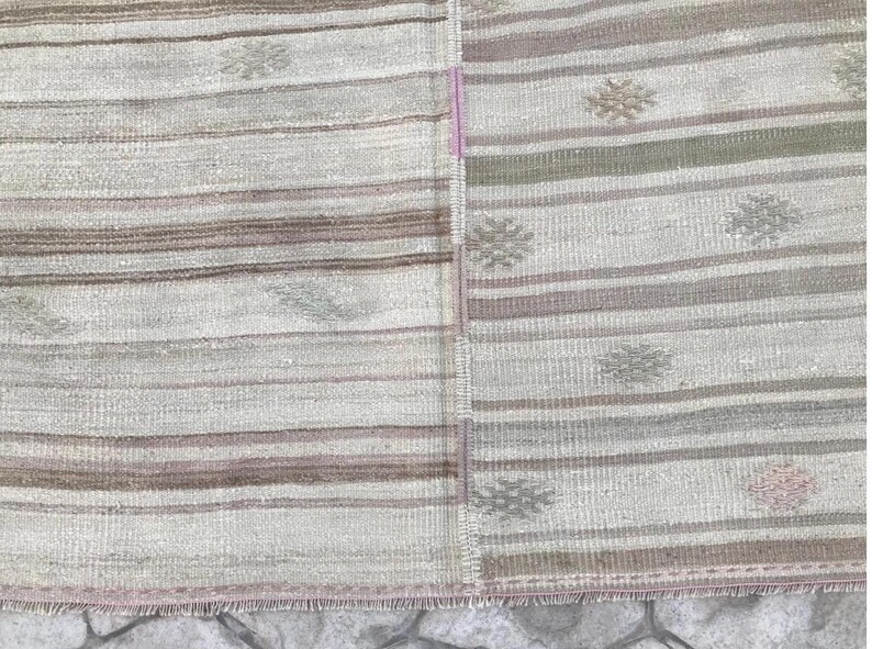 May include: A rectangular, woven rug with a patchwork design. The rug features horizontal stripes in shades of beige, brown, and gray. Floral-like patterns are scattered across the surface. The edges have a fringed border.