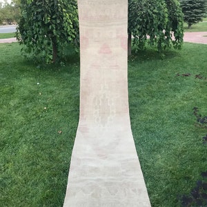 May include: A long, narrow, off-white rug with a subtle pink and white pattern. The rug has a fringed edge and is hanging on a clothesline.