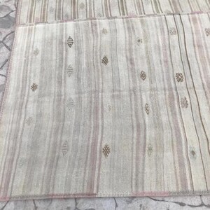 May include: A rectangular, woven rug with a striped pattern in shades of beige, gray, and pale pink. The rug features small, repeating geometric designs in brown. The edges are finished with a pink border, creating a neutral and textured aesthetic.
