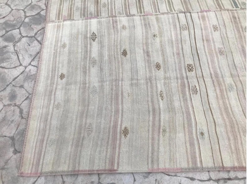 May include: A rectangular, woven rug with a striped pattern in shades of beige, gray, and pale pink. The rug features small, repeating geometric designs in brown. The edges are finished with a pink border, creating a neutral and textured aesthetic.