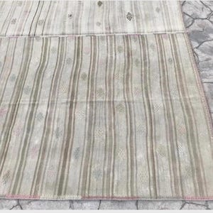 May include: A rectangular, hand-woven rug with a striped pattern in shades of beige, gray, and pink. The rug features vertical stripes and small, decorative floral motifs. The edges are finished with a pink border.