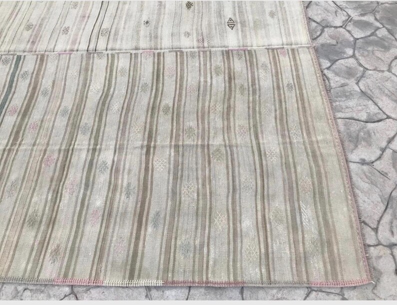 May include: A rectangular, hand-woven rug with a striped pattern in shades of beige, gray, and pink. The rug features vertical stripes and small, decorative floral motifs. The edges are finished with a pink border.