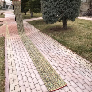 Pode incluir: Um tapete corredor longo e estreito com fundo marrom e intrincados padrões florais e geométricos em tons de verde, amarelo e azul. O tapete tem uma borda vermelha e é exibido ao ar livre em uma calçada de tijolos.