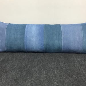 Pode incluir: Uma almofada longa e retangular com aparência de ganga. Apresenta painéis verticais em tons de azul variados, criando um efeito listrado. A almofada é projetada para uso decorativo ou funcional.