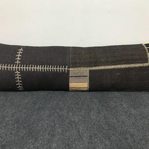 Puede incluir: Un cojín largo y rectangular de tejido gris oscuro. Presenta un diseño de cruz cosida con hilo color crema en un extremo y parches geométricos en tonos neutros. El cojín es una mezcla de texturas y patrones.