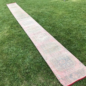 May include: A long, rectangular rug with a pink, white, and gray pattern. The rug has a fringe border and is laid out on a green lawn.