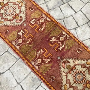May include: A vintage Turkish runner rug with a red, orange, and green geometric pattern. The rug has a fringe on both ends and is made of wool.