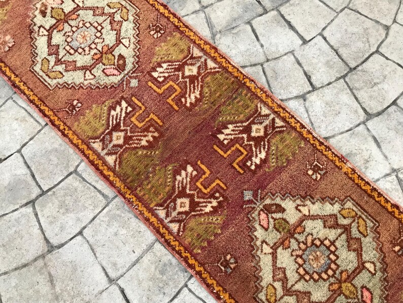 May include: A vintage Turkish runner rug with a red, orange, and green geometric pattern. The rug has a fringe on both ends and is made of wool.