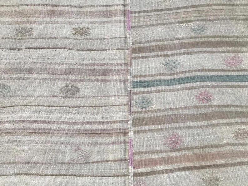 May include: A close-up of a section of a rug with a striped pattern in shades of gray, brown, and pink. The rug has small, repeating floral-like designs. The texture of the rug is visible, suggesting a woven construction.