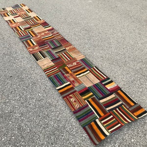 May include: A long, rectangular patchwork rug with a variety of colours and patterns. The rug is made of different fabrics sewn together in a patchwork style. The rug is made up of squares and rectangles of different sizes and colours. The rug is a mix of brown, red, orange, yellow, green, blue, and black.