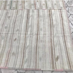 May include: A rectangular rug with a striped pattern in shades of beige, gray, and pink. The rug features vertical stripes of varying widths and small floral motifs. The edges are finished with a pink border. The rug is placed on a stone surface.