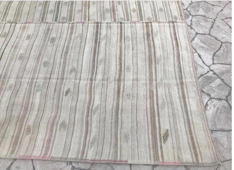May include: A rectangular rug with a striped pattern in shades of beige, gray, and pink. The rug features vertical stripes of varying widths and small floral motifs. The edges are finished with a pink border. The rug is placed on a stone surface.