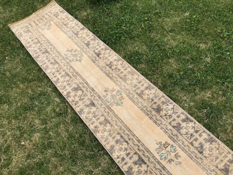 Pu&ograve; includere: Un lungo tappeto da corridoio beige con un bordo a motivi marroni e blu. Il tappeto ha un aspetto sbiadito e vintage.
