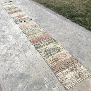 May include: A long, narrow, multi-colored rug with a patchwork design. The rug is made of different fabrics and has a variety of patterns, including floral, geometric, and abstract designs. The rug is laid out on a gray concrete walkway.
