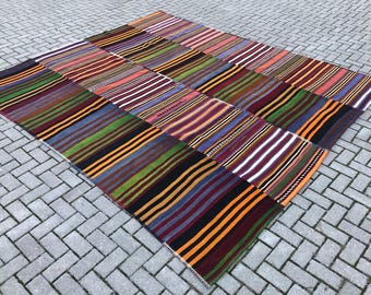 May include: A patchwork rug made of different colored striped fabric. The rug is made up of 16 squares, each with a different color combination of stripes. The rug is laid out on a gray brick floor.