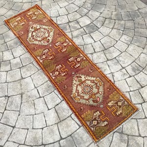 May include: A long, narrow rug with a traditional Turkish design. The rug is red, orange, and cream with a geometric pattern.