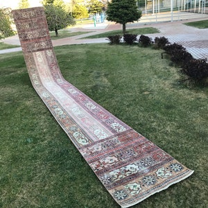 May include: A long, rectangular rug with a cream background and intricate floral and geometric patterns in shades of red, blue, and green. The rug is laid out on a grassy lawn.