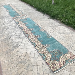 Puede incluir: Una alfombra larga de color azul verdoso con un patrón floral en tonos crema, rosa y marrón. La alfombra está colocada en un patio de concreto gris.