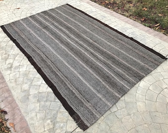 May include: A rectangular, striped rug with a dark brown border. The rug features alternating stripes of dark brown, light brown, and off-white. The rug is made of woven material and is placed on a concrete surface.