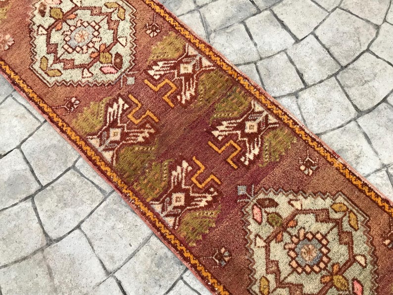 May include: A vintage Turkish runner rug with a red, orange, and green geometric pattern. The rug has a fringe on both ends and is made of wool.