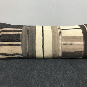 May include: Rectangular decorative pillow with a patchwork design. The pillow features a mix of neutral colors, including shades of brown, beige, and gray, arranged in vertical stripes and rectangular blocks. The pillow is resting on a dark gray surface.