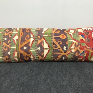 May include: A long, rectangular cushion with a vibrant, patterned design. The cushion displays a patchwork of geometric and abstract shapes in red, green, brown, and cream. The cushion is designed for decorative use.
