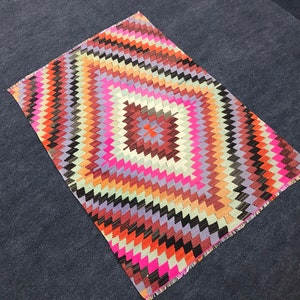 Puede incluir: Una alfombra geométrica colorida con un patrón de diamante en tonos de rosa, morado, naranja, marrón y negro. La alfombra tiene un borde con flecos.
