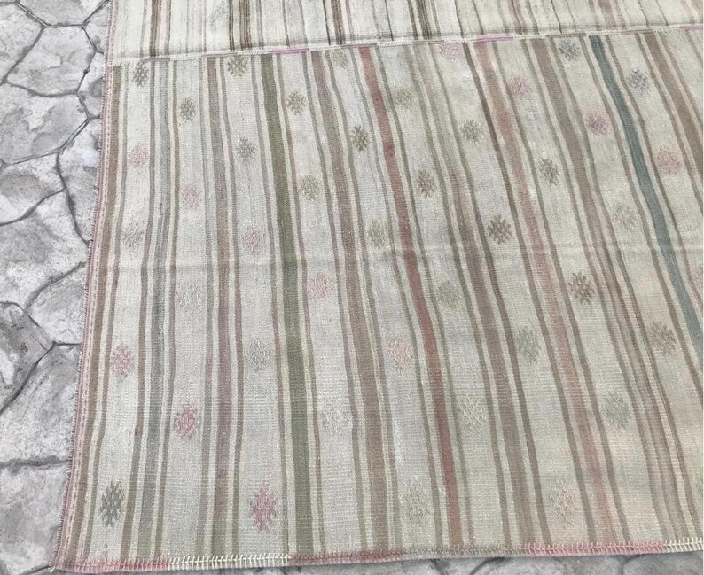 May include: A rectangular rug with vertical stripes in shades of beige, pink, green, and gray. The rug has a faded, vintage appearance with small floral-like patterns scattered throughout. The edges are finished with a pink border.