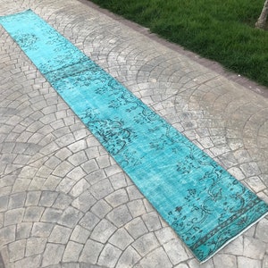 May include: A long, teal blue runner rug with a faded, black floral pattern. The rug is laid out on a gray, patterned concrete surface.