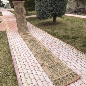 May include: A long, rectangular beige runner rug with an intricate geometric pattern. The rug features a repeating design of squares and floral motifs in shades of brown and green. The edges are finished with a red border.