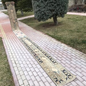 May include: A long, narrow, cream-coloured rug with a black and white geometric pattern along the edges. The rug is laid out on a brick pathway.