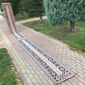 Könnte beinhalten: Ein langer, schmaler Teppich mit einem geometrischen Muster in Rosa und Blau. Der Teppich liegt auf einem gepflasterten Weg in einem Park.