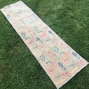 May include: A rectangular beige runner rug with a floral pattern in pink and blue. The rug is placed on green grass and has a faded, vintage appearance. The rug's dimensions are not visible.