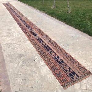 May include: A long, narrow runner rug featuring a repeating geometric pattern in shades of deep navy blue, terracotta orange, and muted cream. The design includes intricate borders and central motifs arranged in a linear, symmetrical composition.