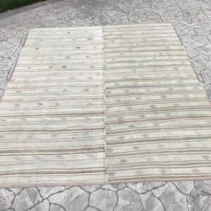 May include: A rectangular, beige rug with horizontal stripes in shades of brown, green, and pink. The rug is divided into two sections and has a pink border. The rug is placed on a stone surface.
