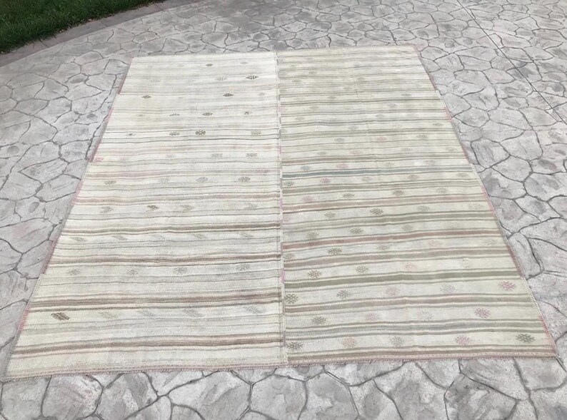 May include: A rectangular, beige rug with horizontal stripes in shades of brown, green, and pink. The rug is divided into two sections and has a pink border. The rug is placed on a stone surface.